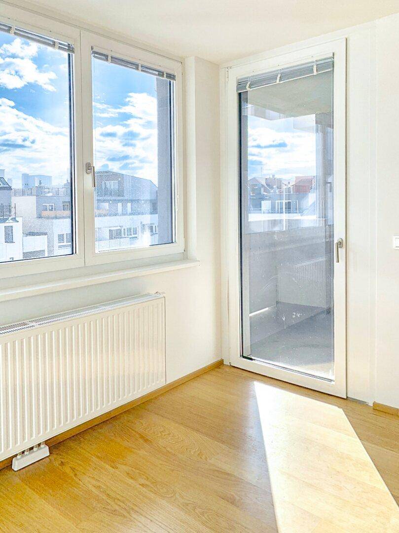 Heller Raum mit großen Fenstern, Heizkörper und Zugang zu einem sonnigen Balkon.