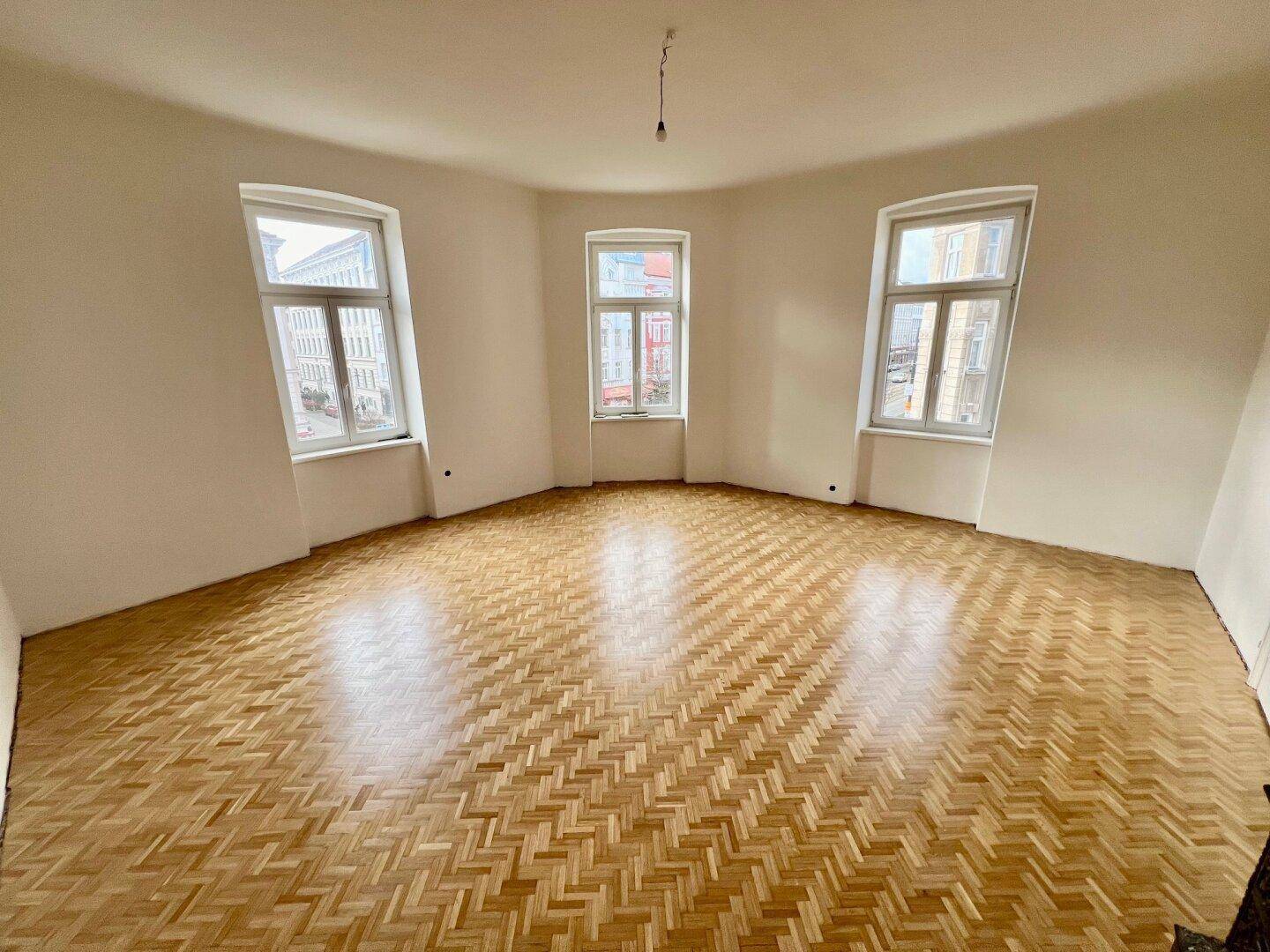 Geräumiges Wohnzimmer mit Fischgrätparkett und drei großen Fenstern für viel Tageslicht.