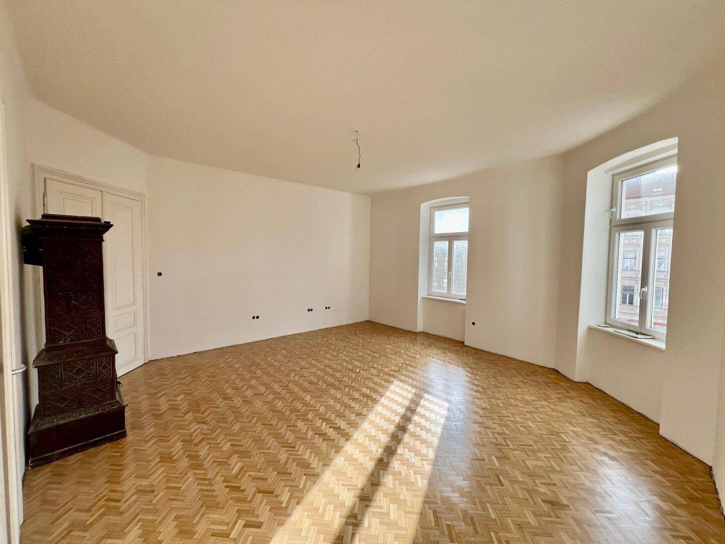Lichtdurchflutetes Zimmer mit Fischgrätparkett, hohen Decken und einem historischen Kachelofen.