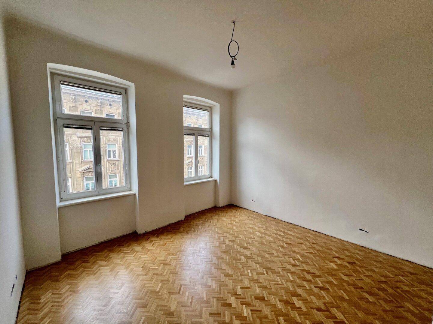 Geräumiges Zimmer mit Fischgrätparkett und zwei großen Fenstern mit Blick auf die Stadt.