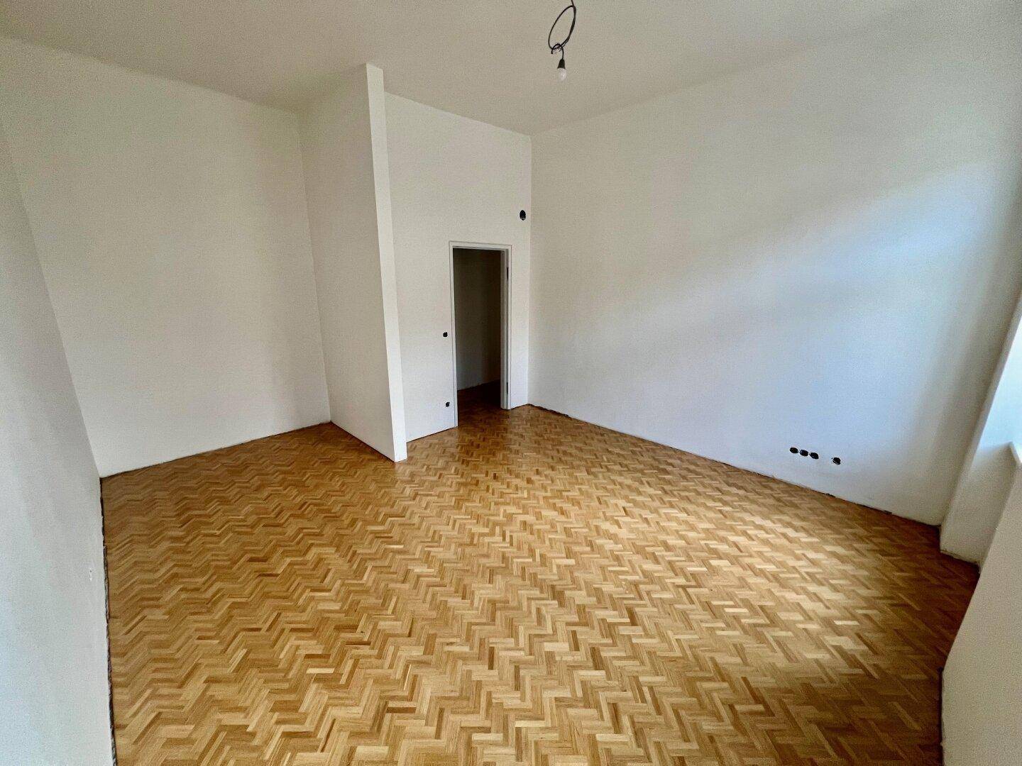 Helles Zimmer mit Fischgrätparkett und einem Durchgang zu einem angrenzenden Raum.