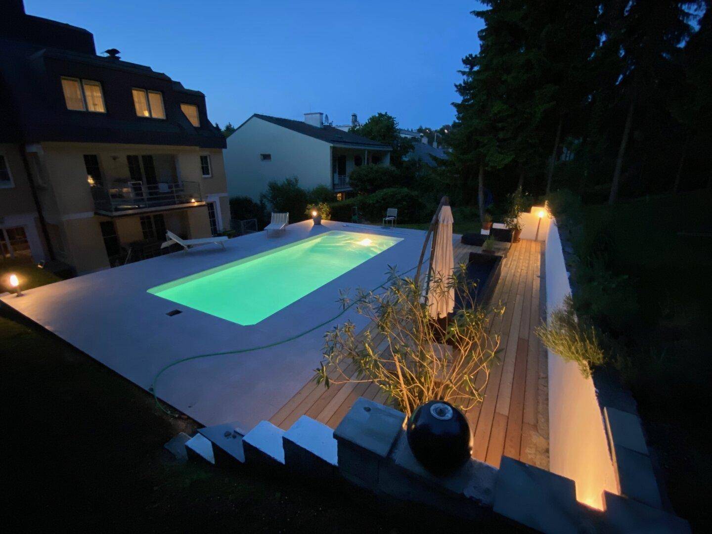 Beleuchteter Gartenbereich mit Swimmingpool und Terrasse bei Nacht.