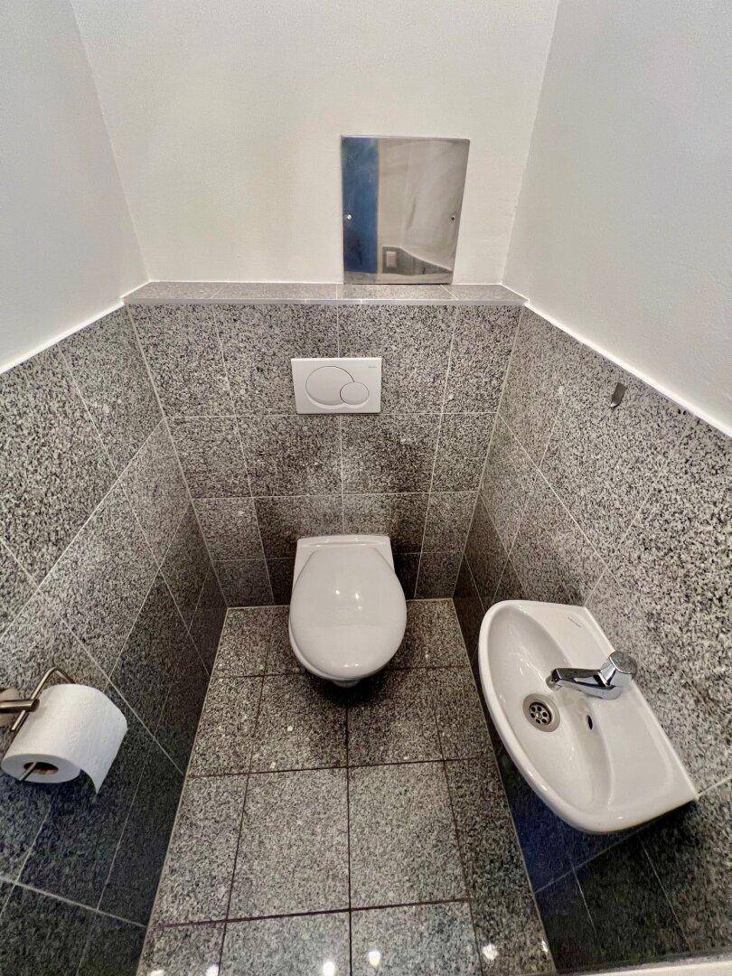 Kleines Gäste-WC mit Wand-WC, Waschbecken und grauen Fliesen.