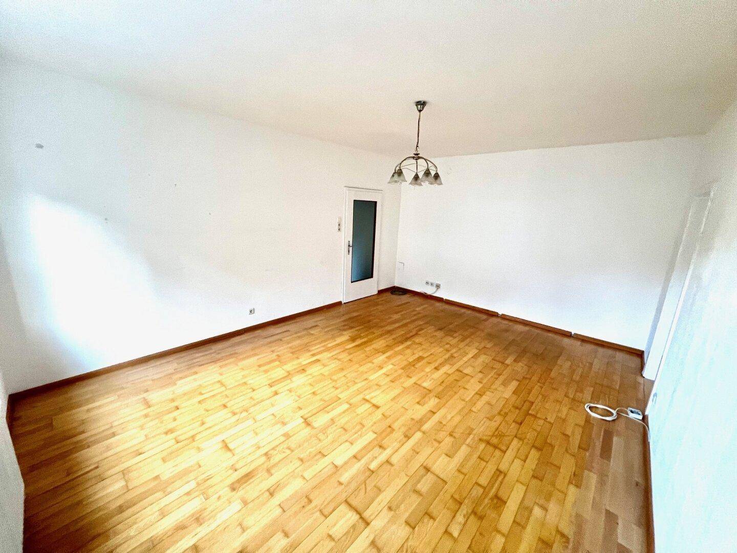 Geräumiges, leeres Zimmer mit hellem Parkettboden und einer Tür, die zu einem anderen Bereich führt.