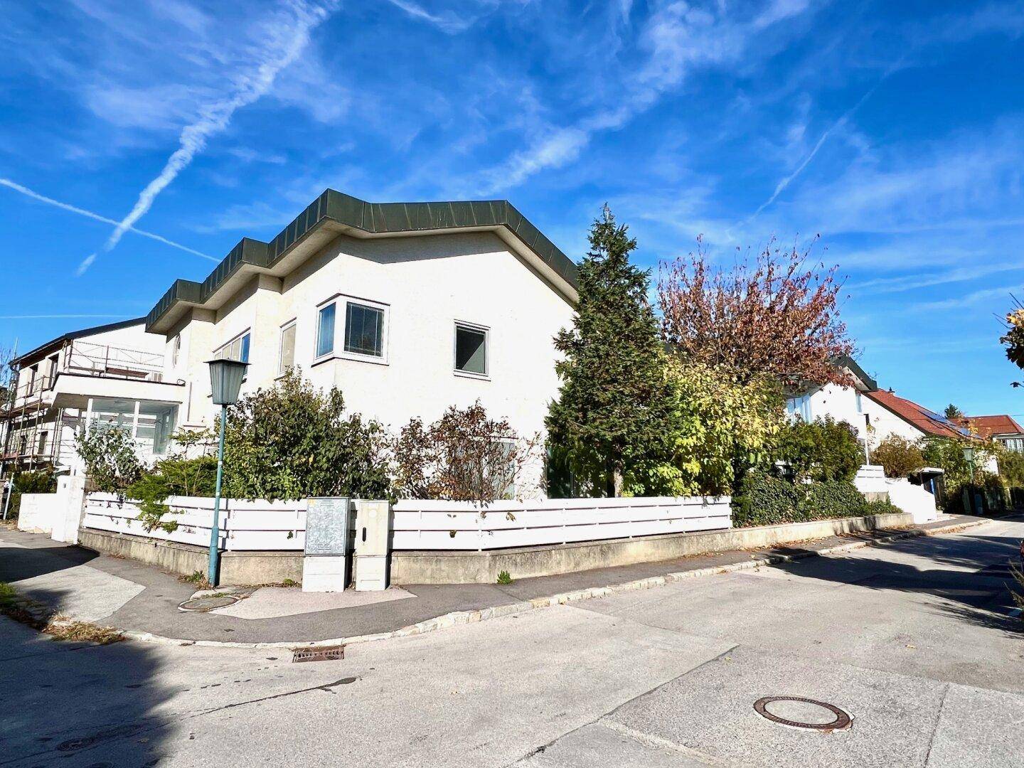 Wohnhaus an einer Straßenecke mit grüner Bepflanzung und klarem Himmel.