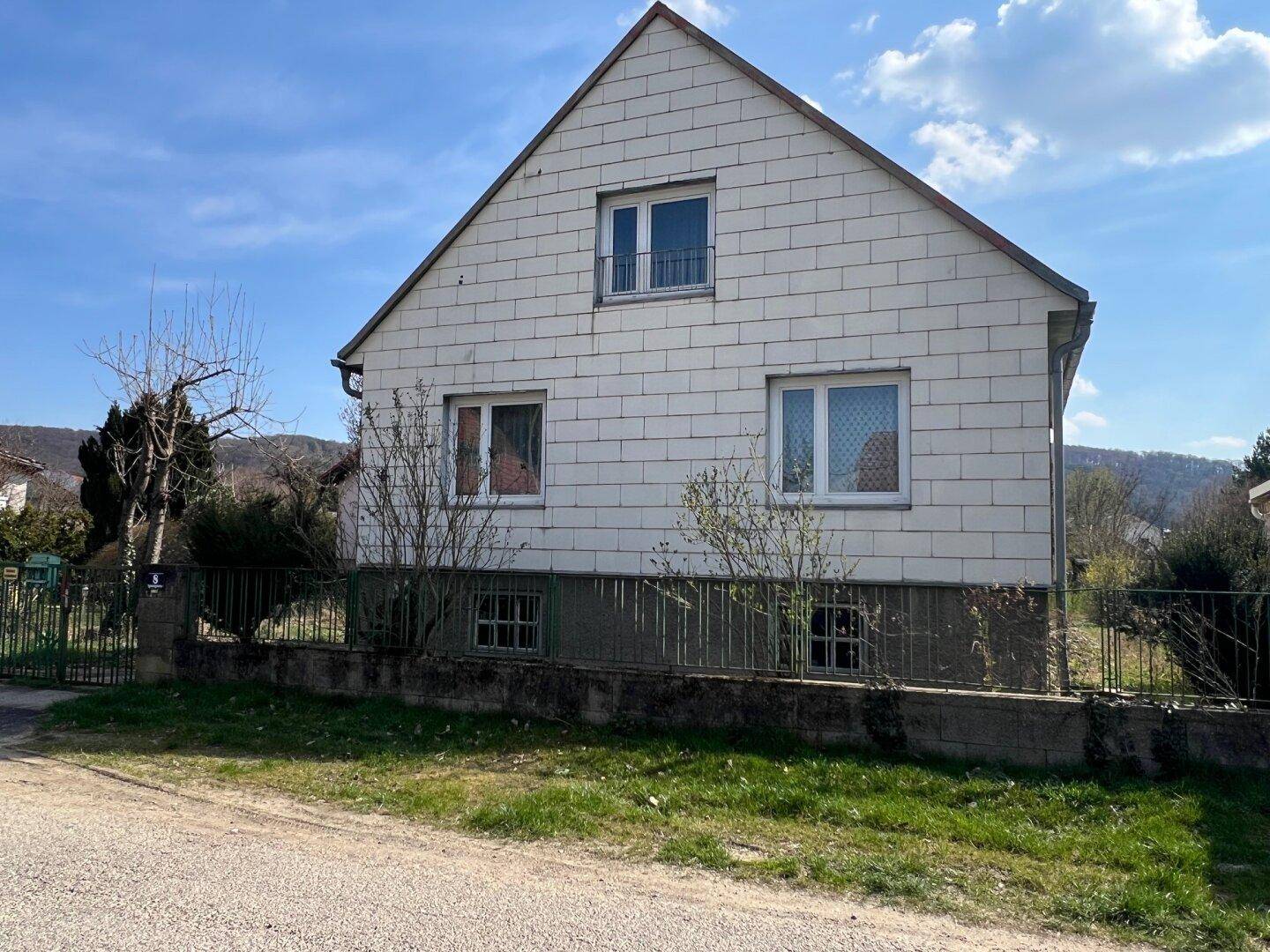 Vorderansicht des Wohnhauses mit weißer Fassade, Satteldach und mehreren Fenstern, umgeben von einem Zaun.