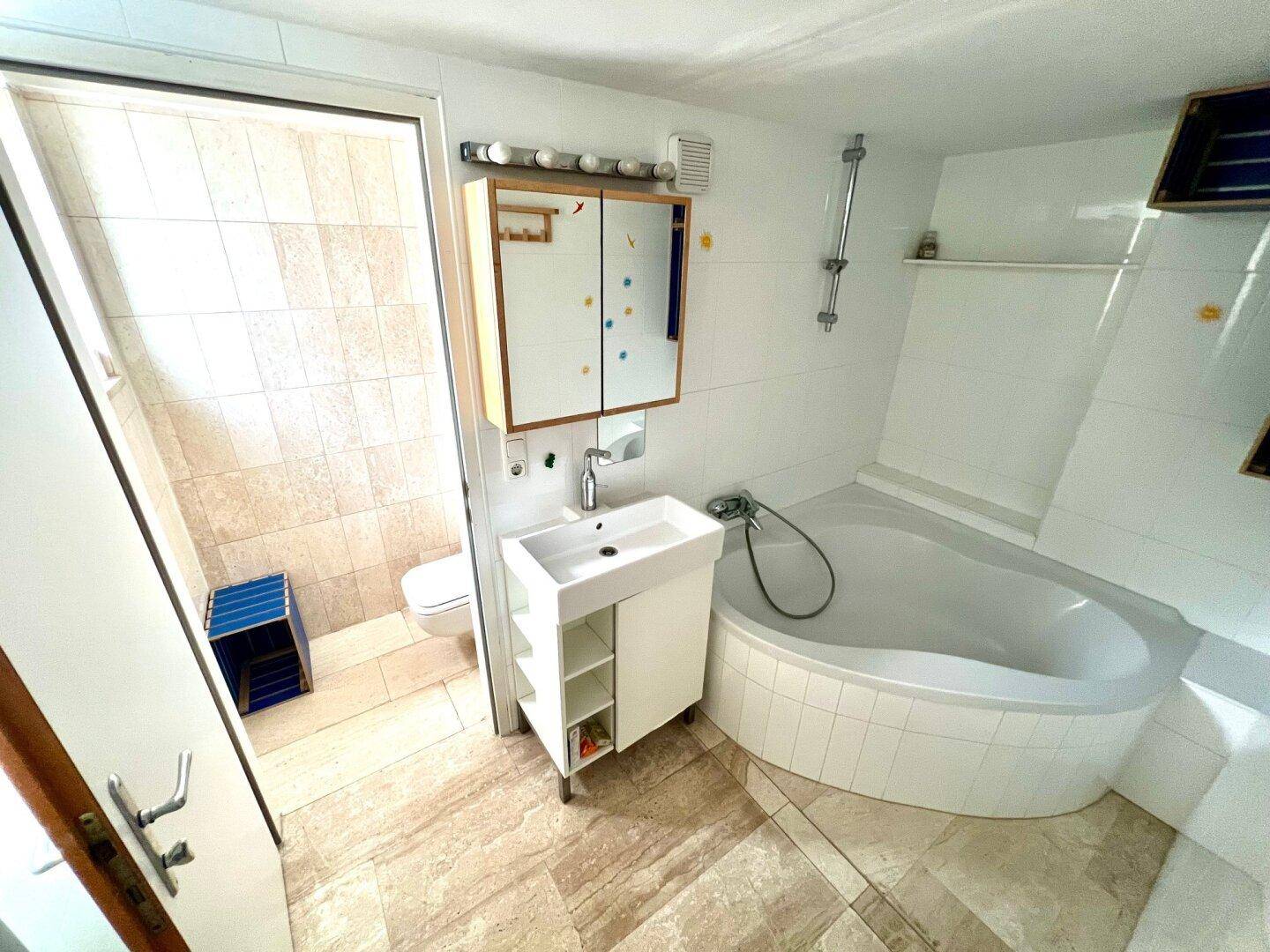 Badezimmer mit Eckbadewanne, modernem Waschbecken und Fliesenboden in hellen Farbtönen.