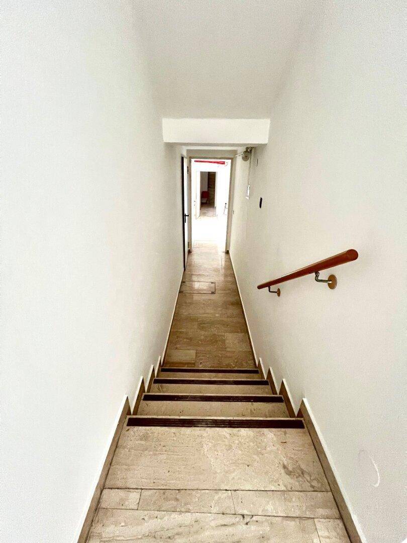 Treppenabgang mit Holzgeländer und hellen Wänden, der in einen unteren Bereich führt.