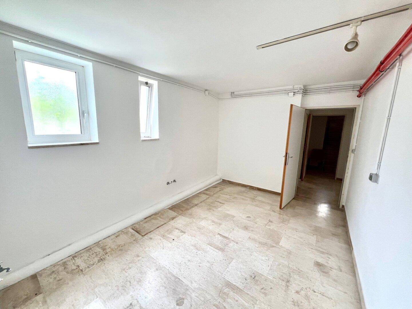 Kellerraum mit Steinboden, zwei Fenstern und sichtbaren Leitungen, funktional gestaltet.