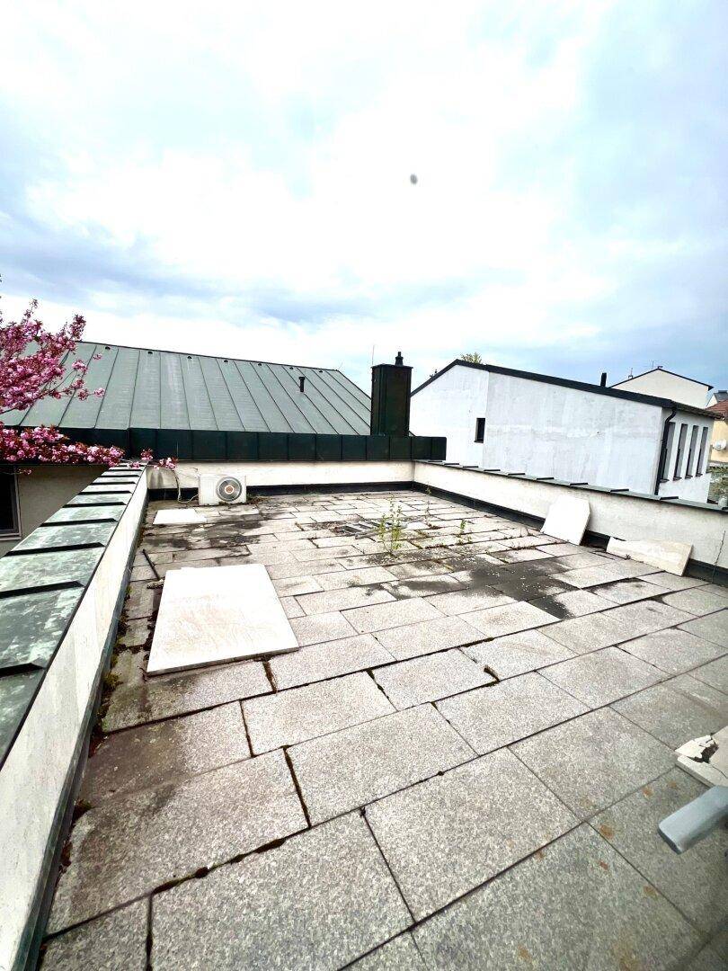 Ungepflegte Dachterrasse mit Steinplatten und Blick auf umliegende Dächer und Bäume.