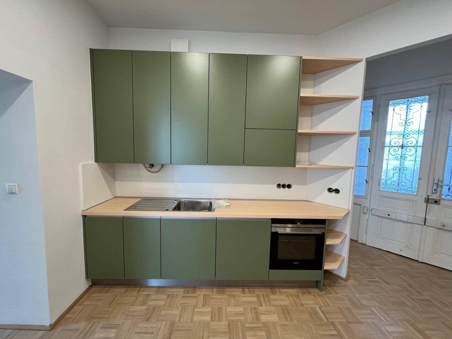 Neue Einbauküche mit grünen Fronten, offenen Holzregalen und integrierter Spüle.