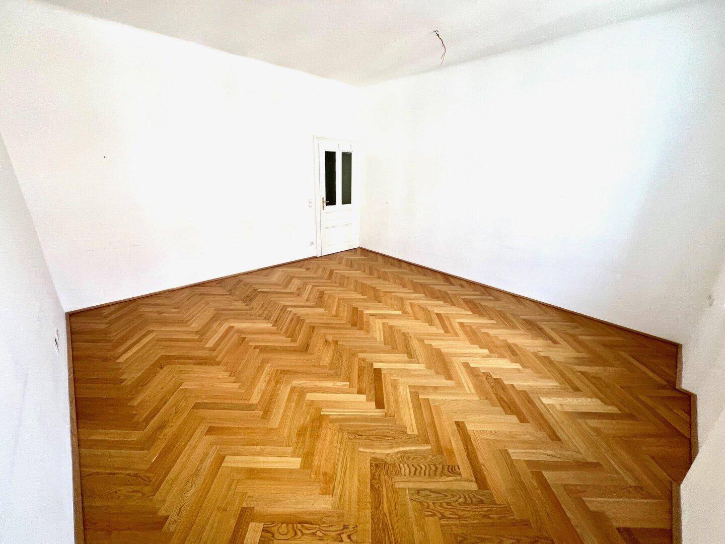 Geräumiges Zimmer mit elegantem Fischgrätparkett und einer weißen Tür, ideal für Wohnzwecke.