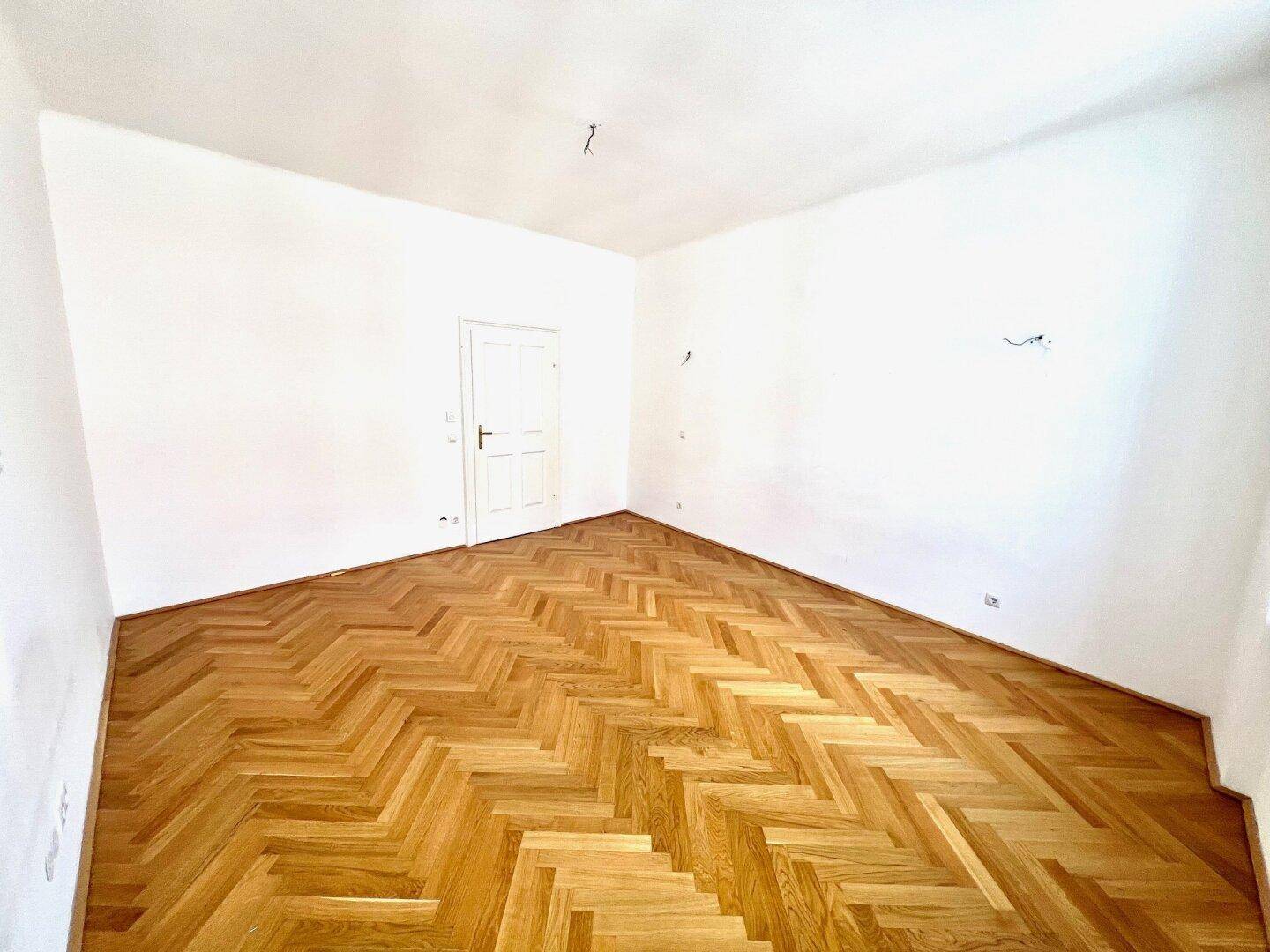 Helles Zimmer mit schönem Fischgrätparkett und einer schlichten weißen Tür.