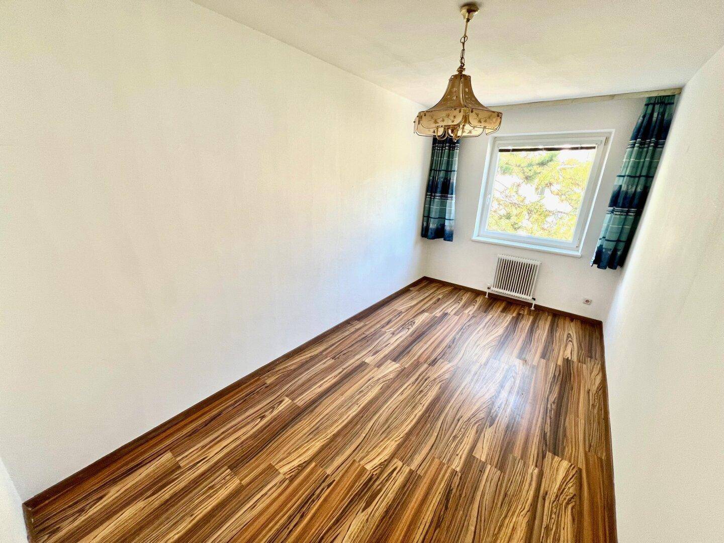 Kleineres Zimmer mit auffälligem Holzboden, einem Fenster und Vorhängen in Blau- und Grüntönen.