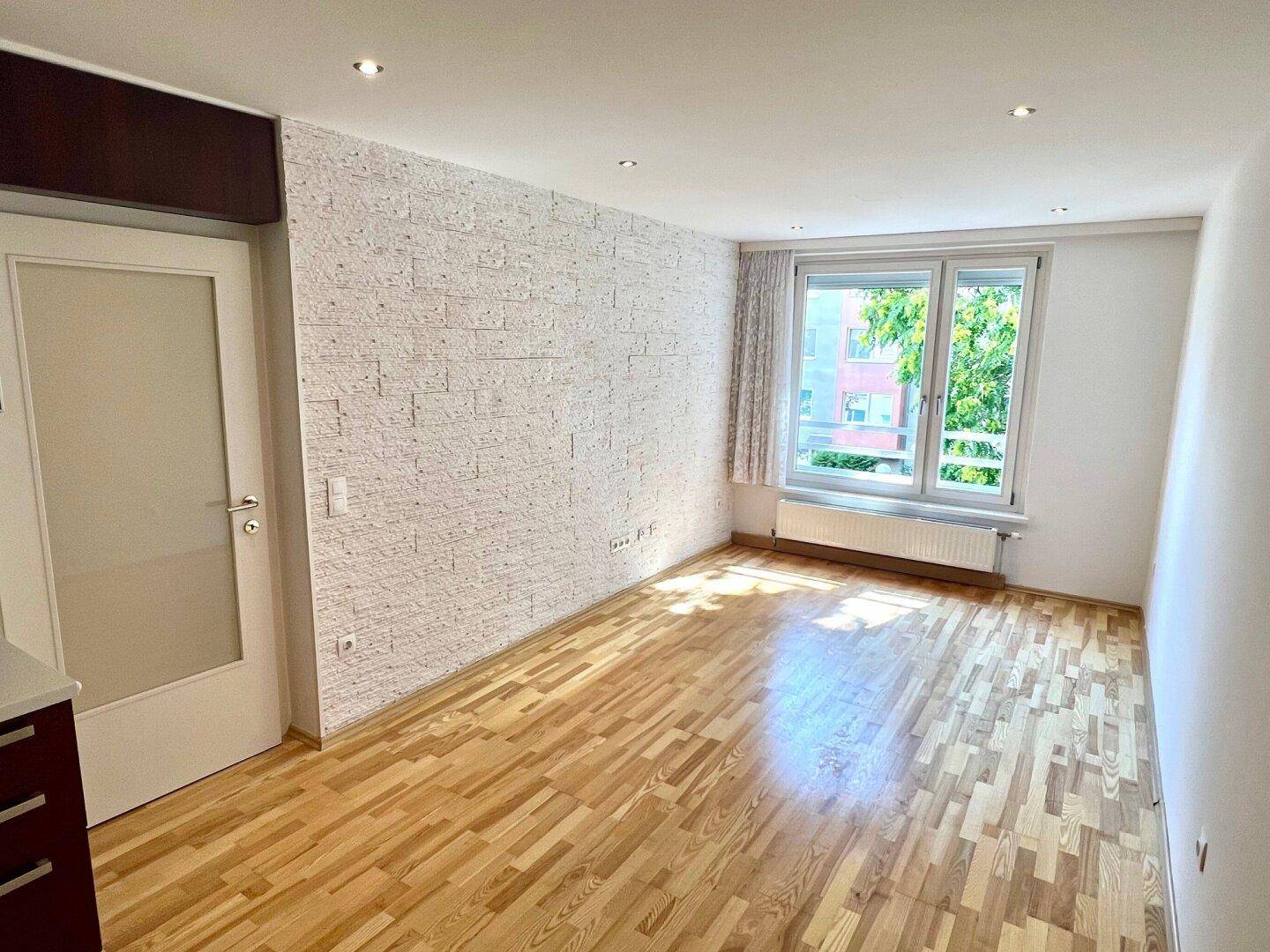 Helles Wohnzimmer mit Parkettboden, einer strukturierten Akzentwand und großem Fenster mit Blick ins Grüne.