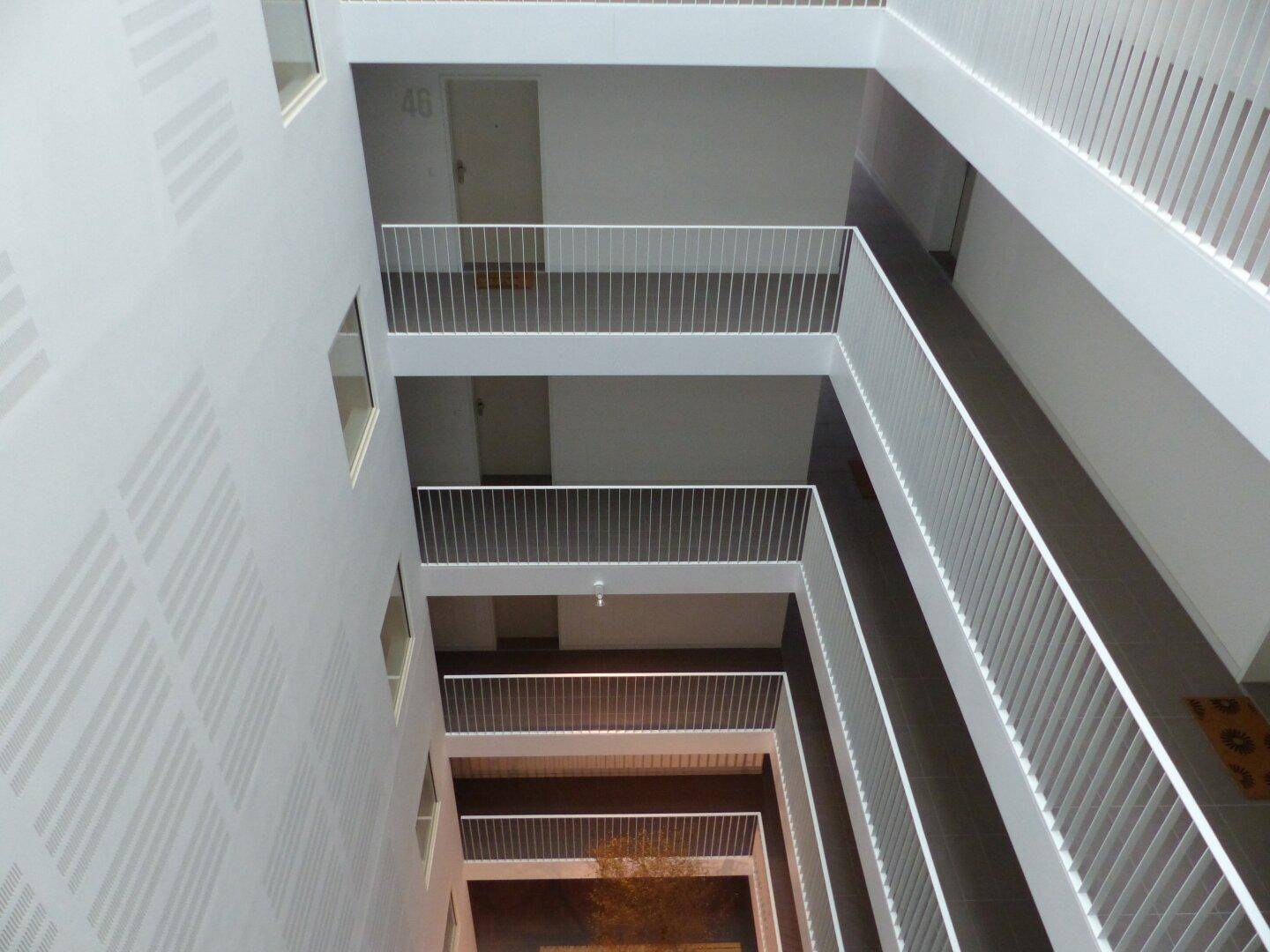 Blick in das helle Atrium eines modernen Wohngebäudes mit mehreren Etagen und Geländern.