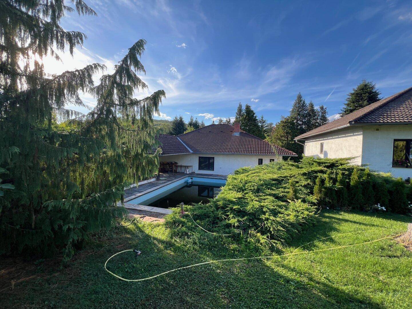 Weitläufiger Gartenbereich mit zwei Wohnhäusern und einem Pool im Hintergrund unter blauem Himmel.