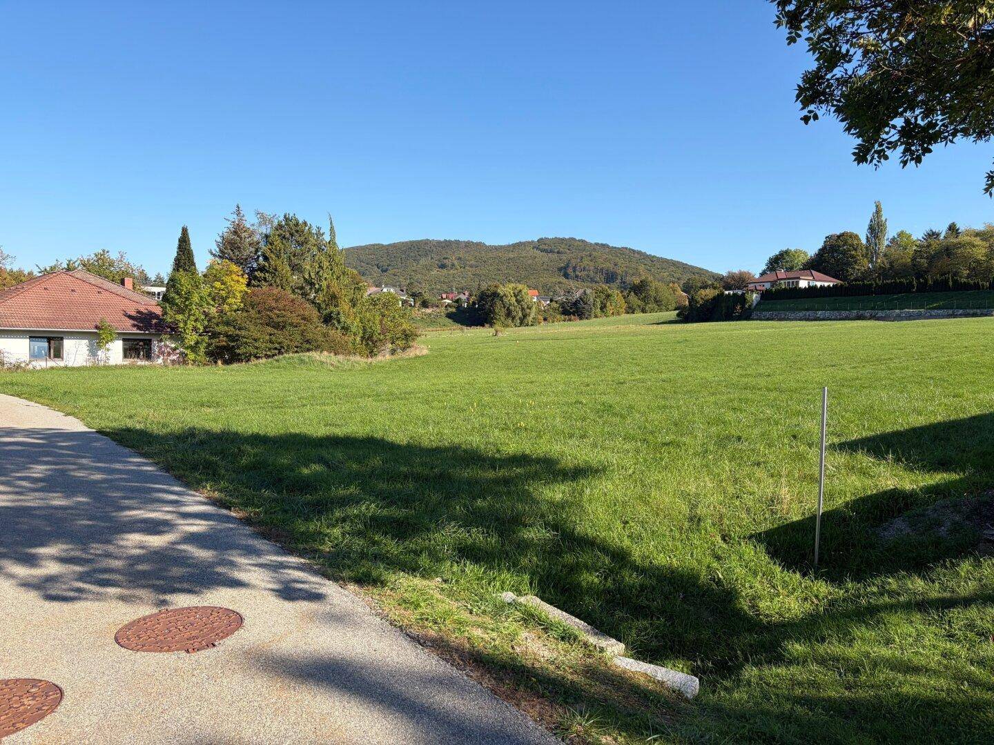 Sonnige Grünfläche mit Blick auf bewaldete Hügel und ländliche Bebauung in der Ferne.
