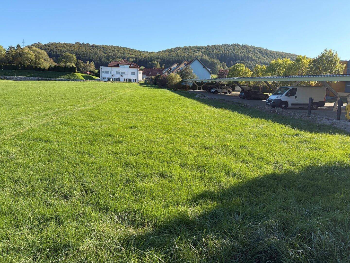 Grüne Wiese mit Blick auf bewaldete Hügel und eine überdachte Parkfläche mit Fahrzeugen am Rand.