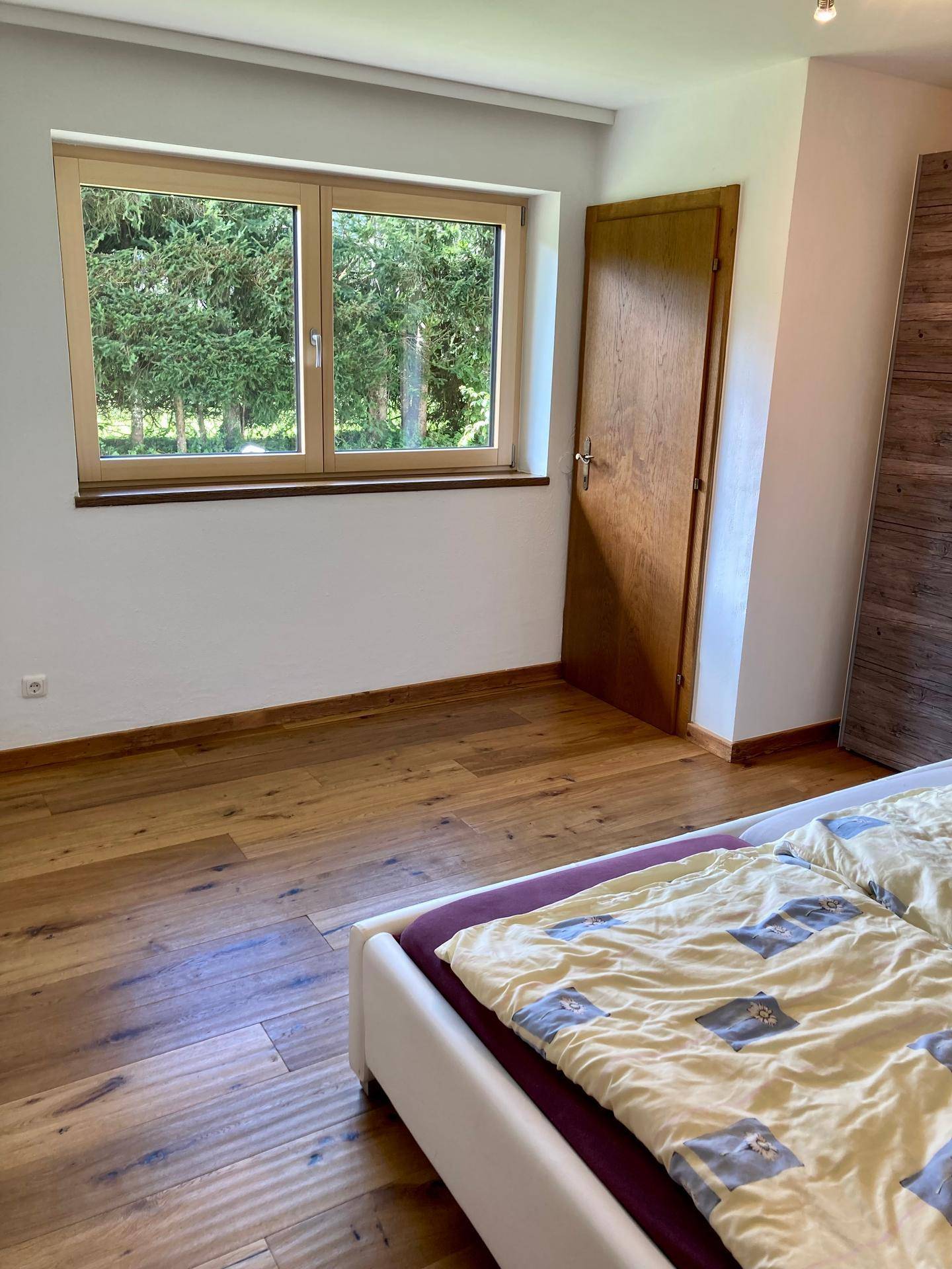 Helles Schlafzimmer mit Holzboden und Fenster mit Blick ins Grüne, schlicht eingerichtet.