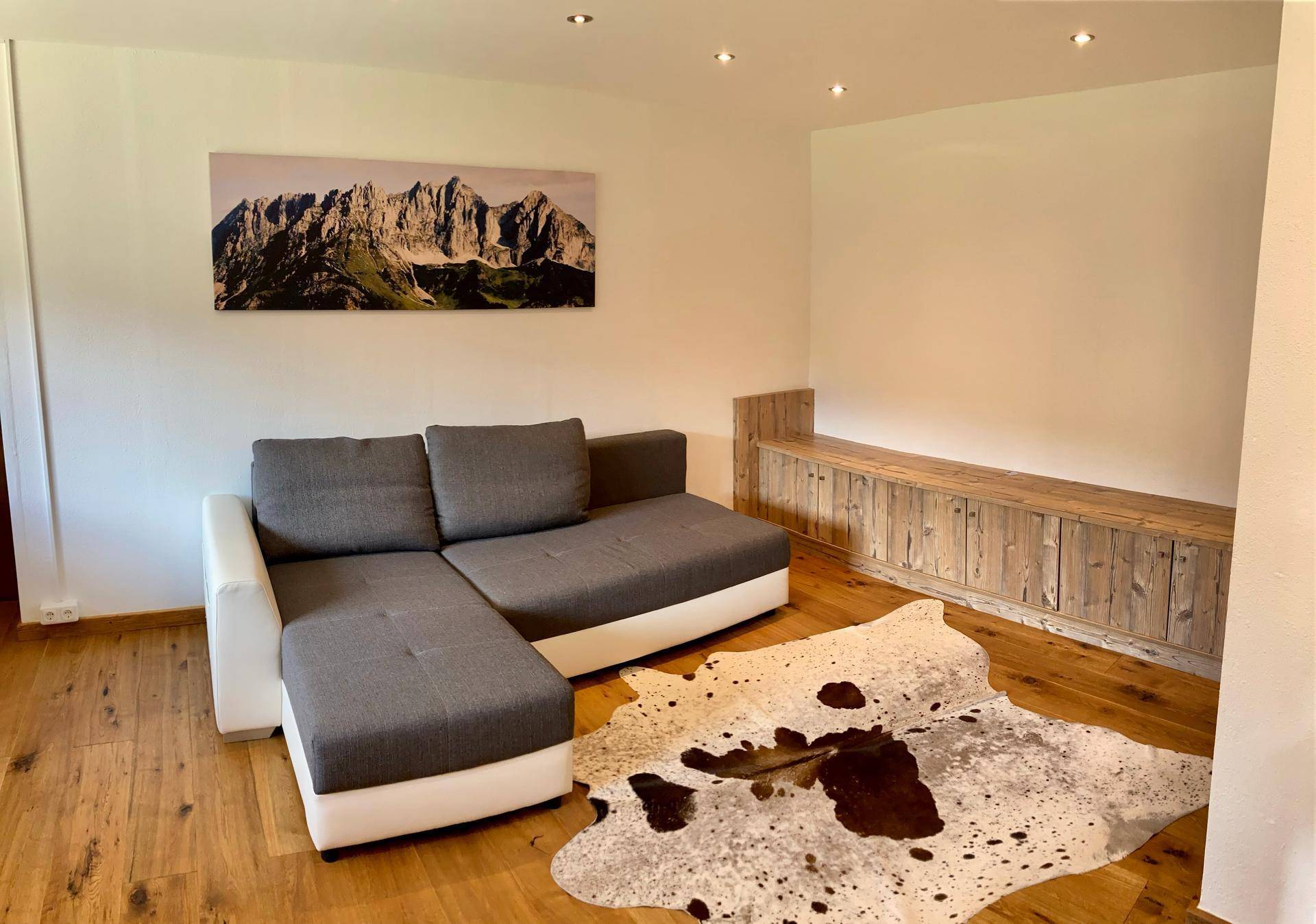 Gemütlicher Wohnbereich mit Ecksofa, Kuhfellteppich und Bergbild an der Wand.