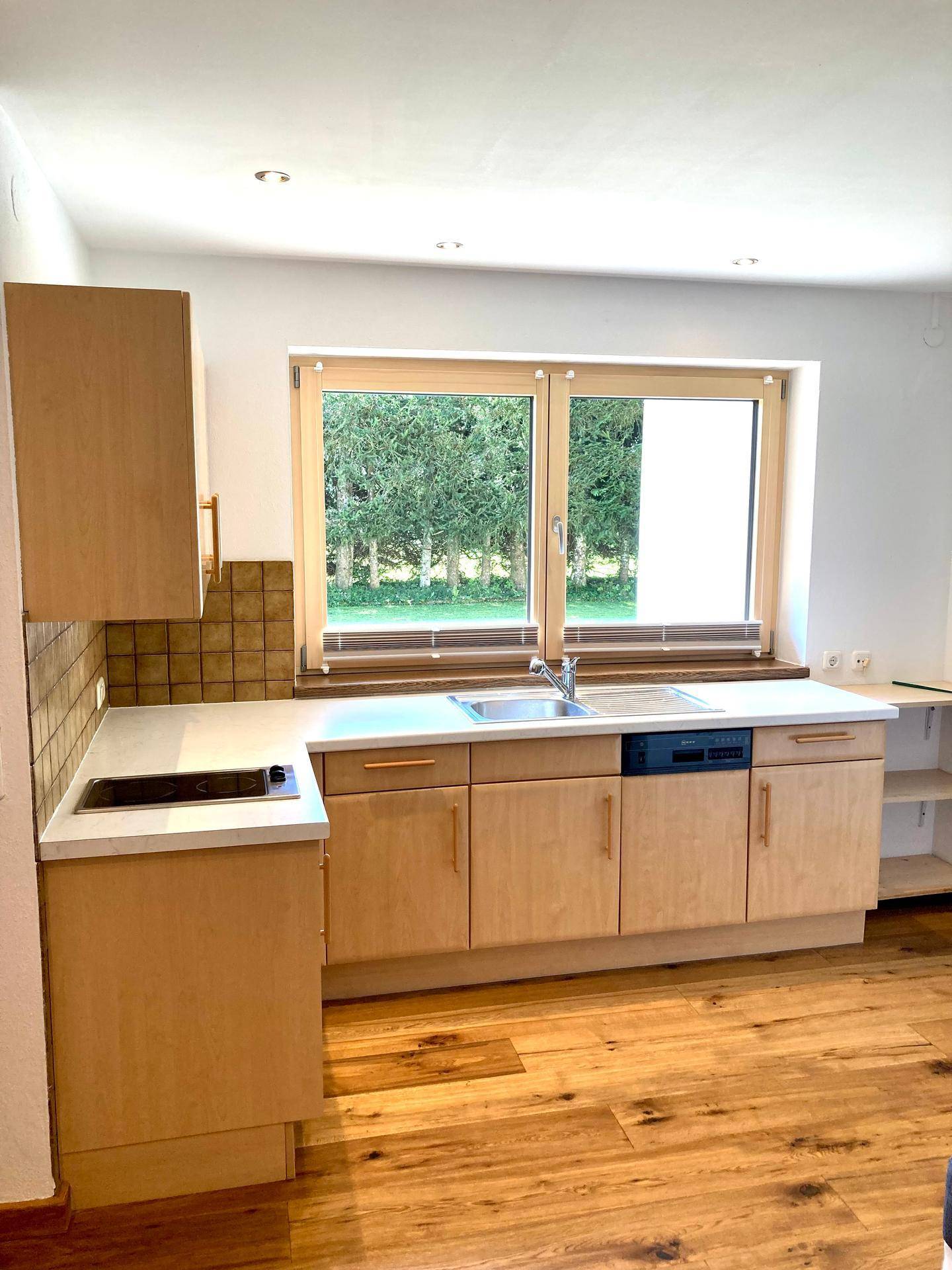 Helle Küchenzeile mit Holzschränken, Spüle und Fenster mit Blick ins Grüne.