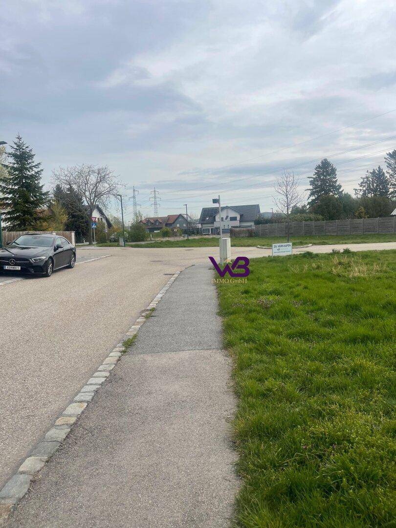 Wohnstraße mit Bürgersteig und Grünstreifen, im Hintergrund sind Häuser und Stromleitungen zu sehen.