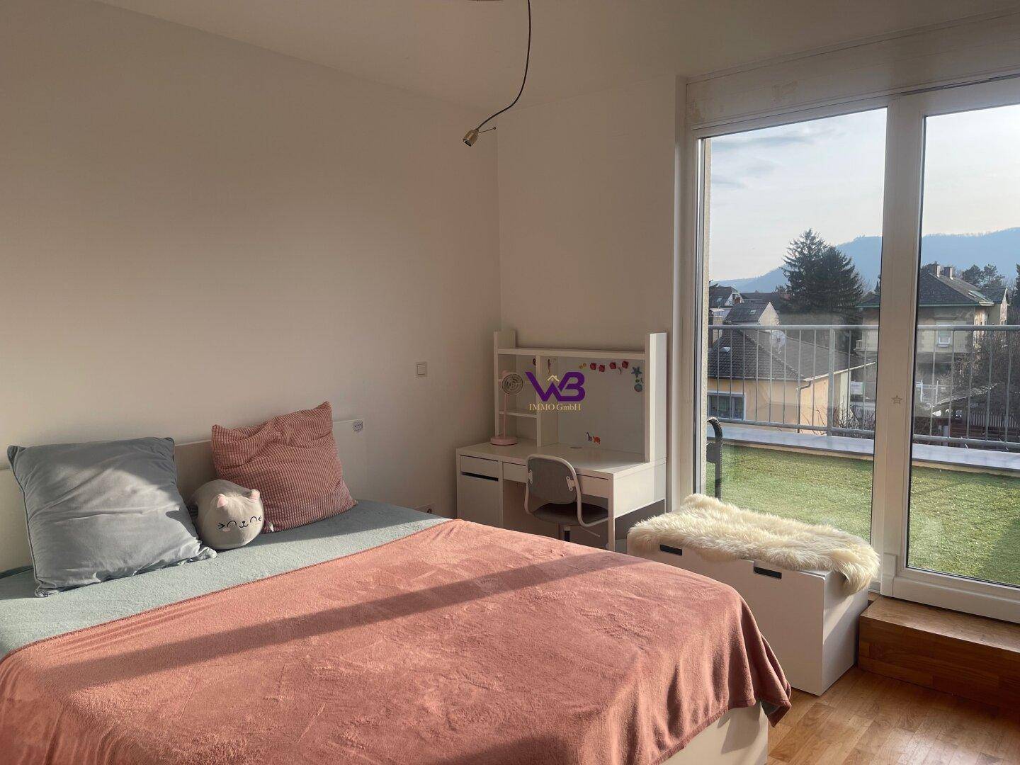 Schlafzimmer mit Bett, Schreibtisch und großen Fenstern zum begrünten Außenbereich.