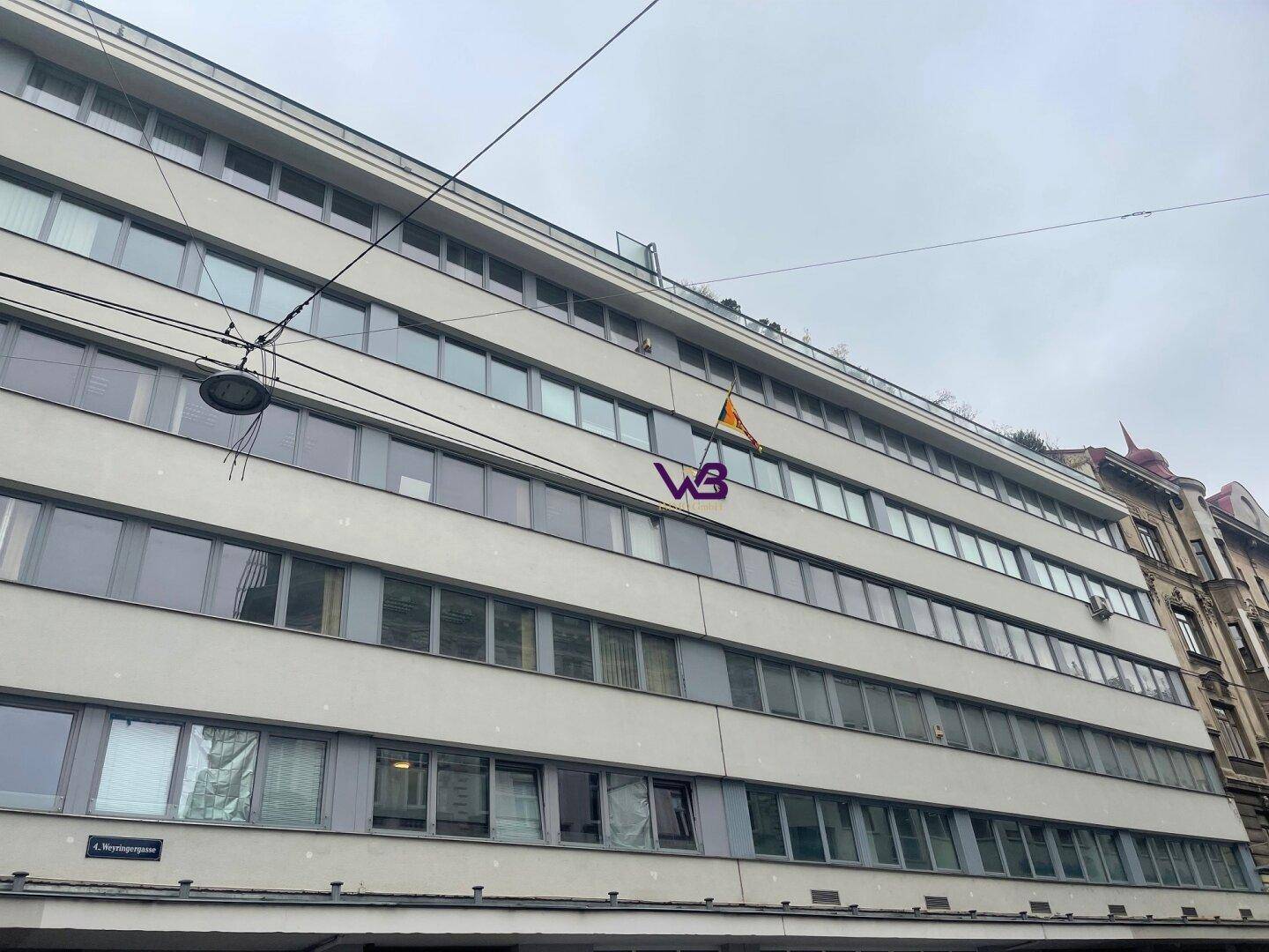 Ansicht der oberen Stockwerke eines Gebäudes mit zahlreichen Fenstern und einer sichtbaren Dachterrasse.