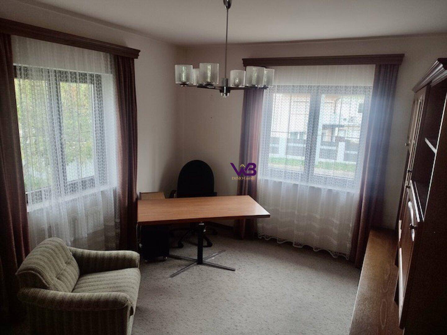 Helles Arbeitszimmer mit Schreibtisch, Sessel und zwei Fenstern, die viel Tageslicht hereinlassen.