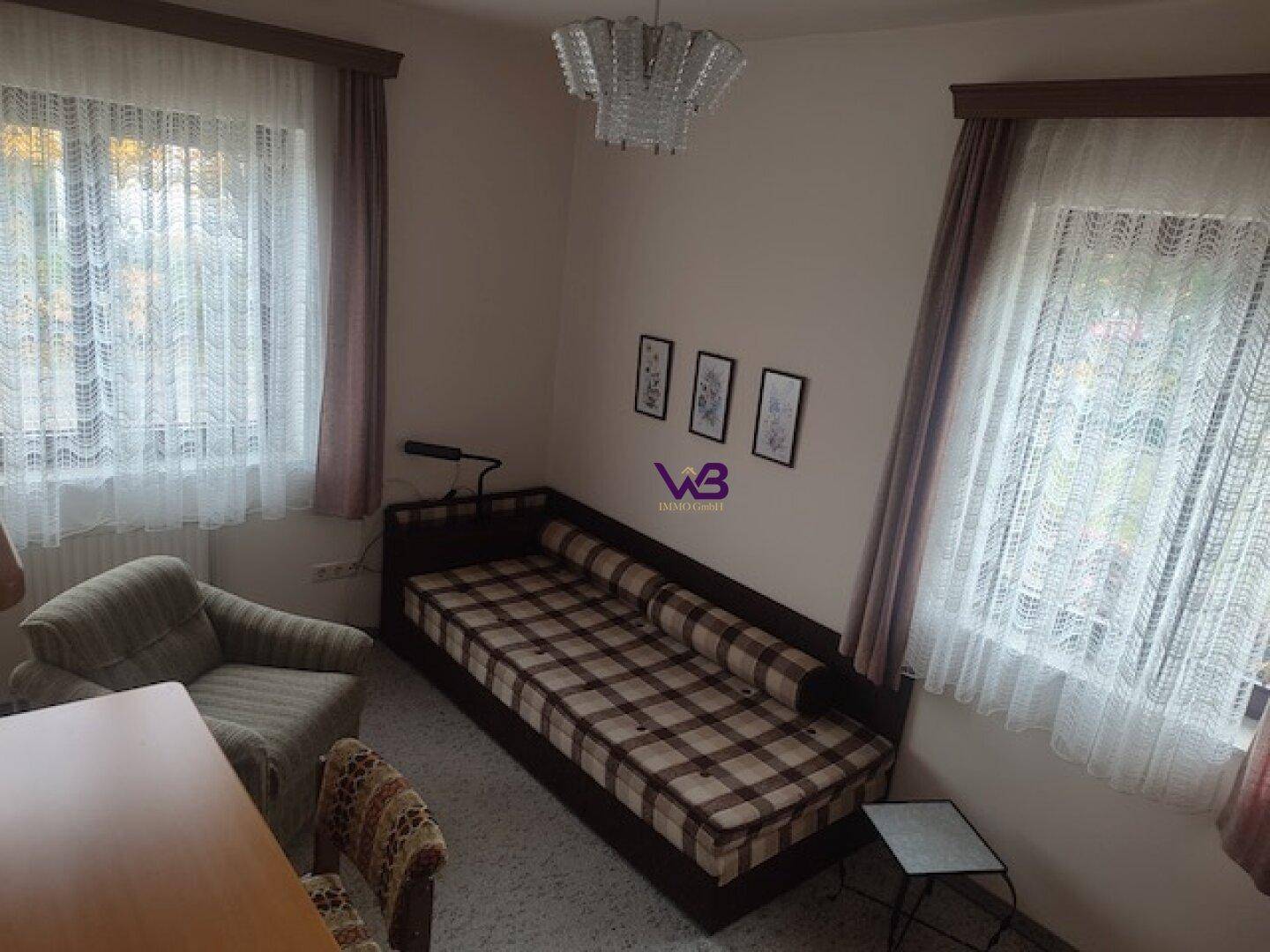 Schlafzimmer mit Tagesbett, Teppichboden und zwei Fenstern mit Vorhängen.