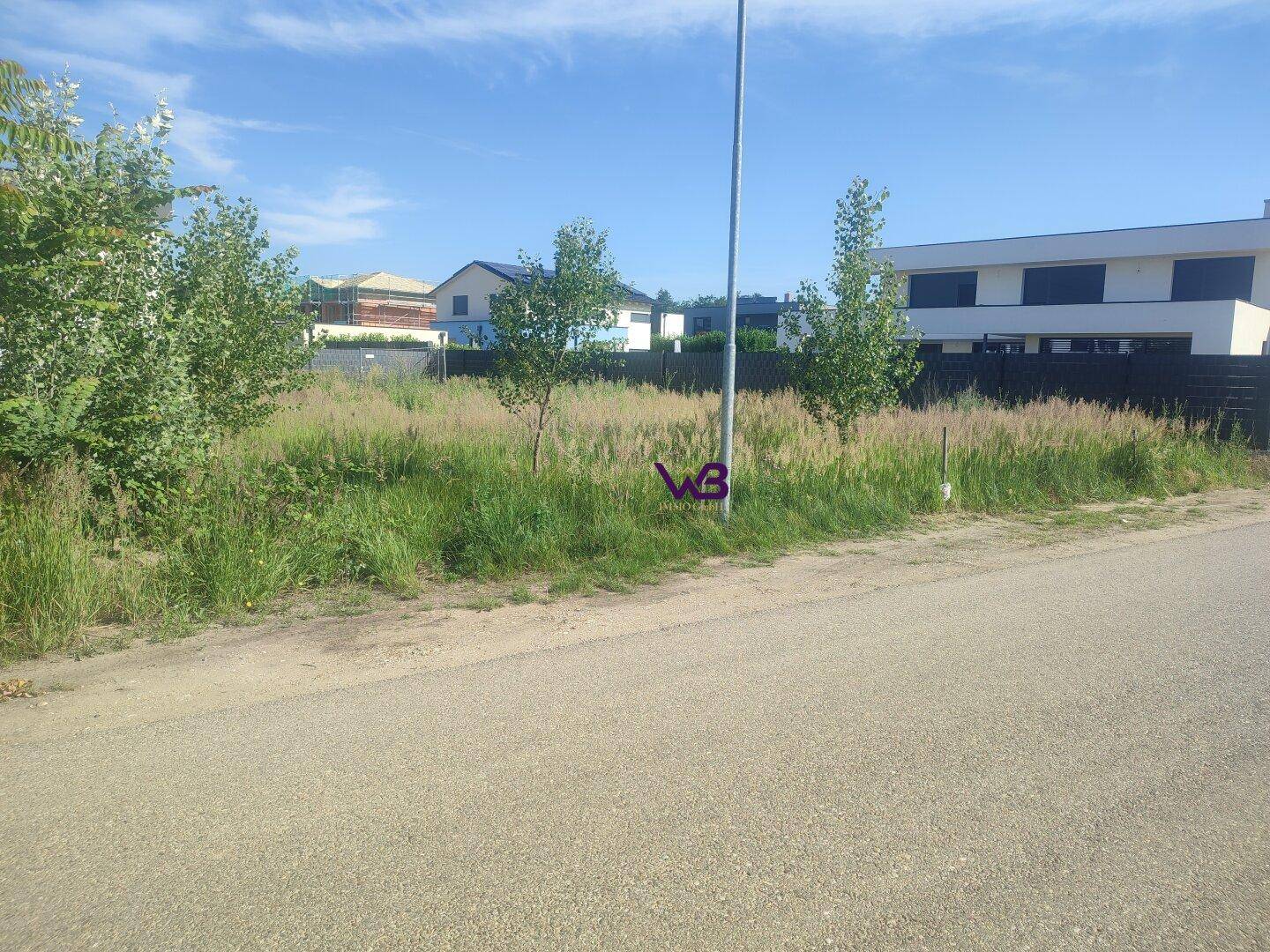 Weitläufiges unbebautes Grundstück mit hohem Gras und modernen Wohnhäusern im Hintergrund.
