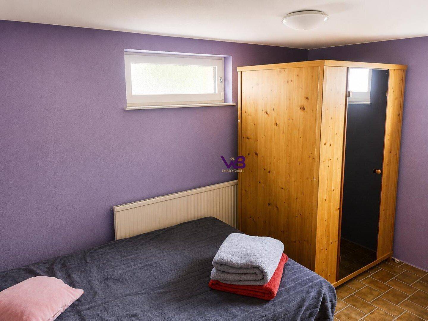 Kellerzimmer mit lila Wänden, Bett und Kleiderschrank, nutzbar als Gästezimmer oder Hobbyraum.