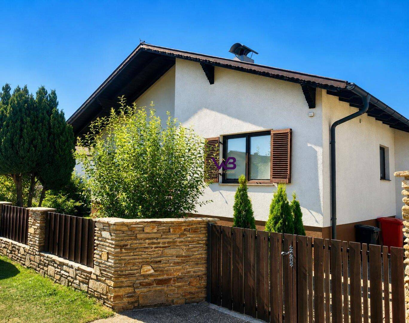 Seitenansicht des Hauses mit traditionellem Holzzaun und Steinmauer, umgeben von Grün.