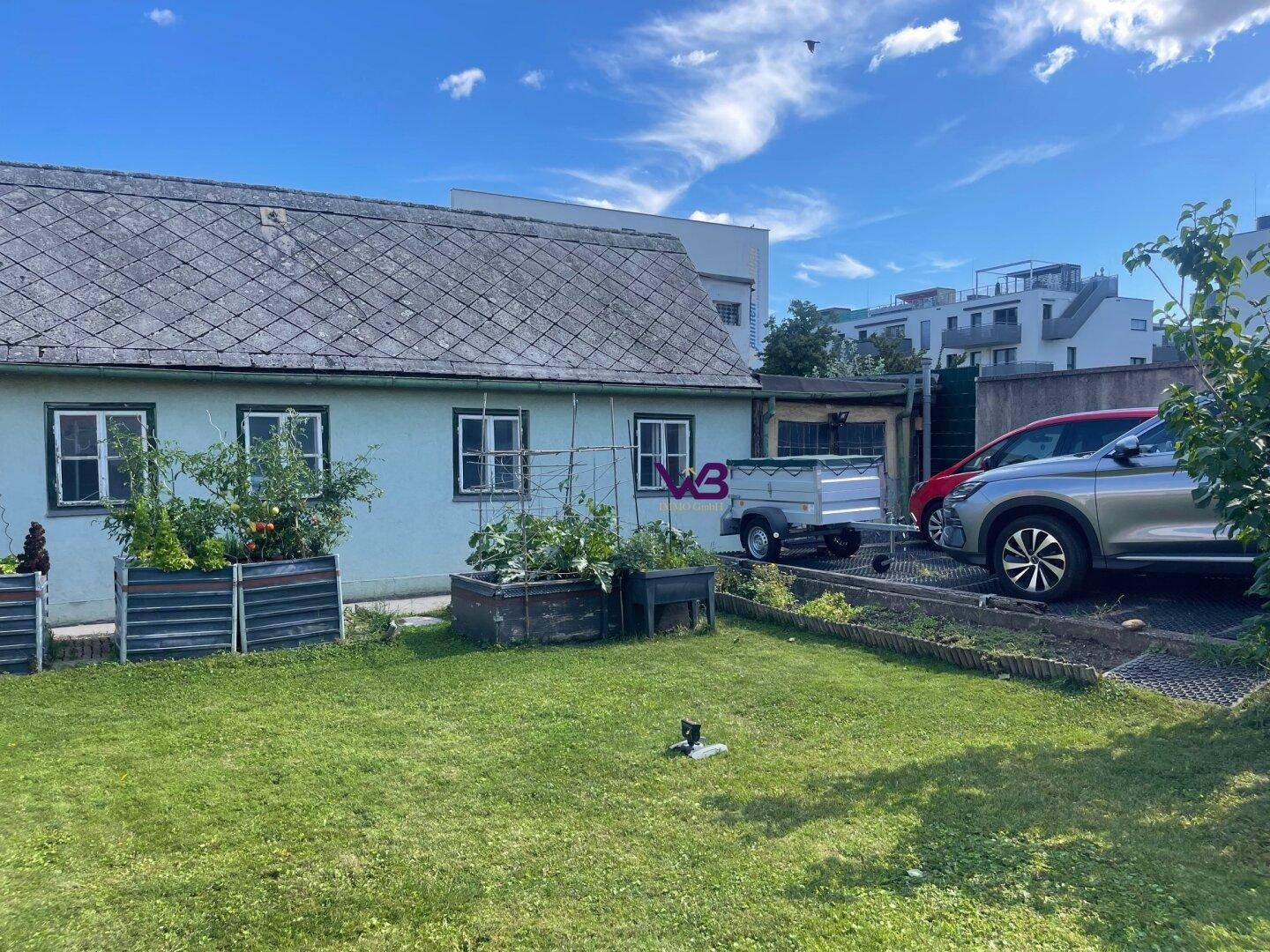Gartenbereich eines älteren Hauses mit Rasen, Hochbeeten und geparkten Autos im Hintergrund.