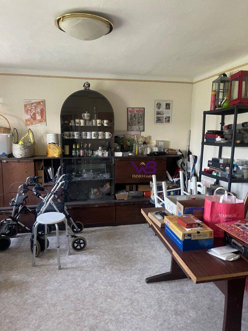 Unaufgeräumter Innenraum mit verschiedenen Möbeln, Kisten und einem Rollator, unter einer Holzdecke.