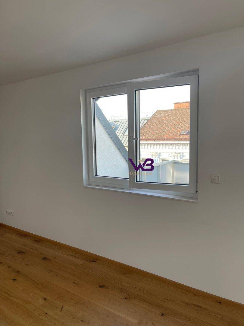 Heller Raum mit Holzboden und einem Fenster, das einen Blick auf die Dächer bietet.