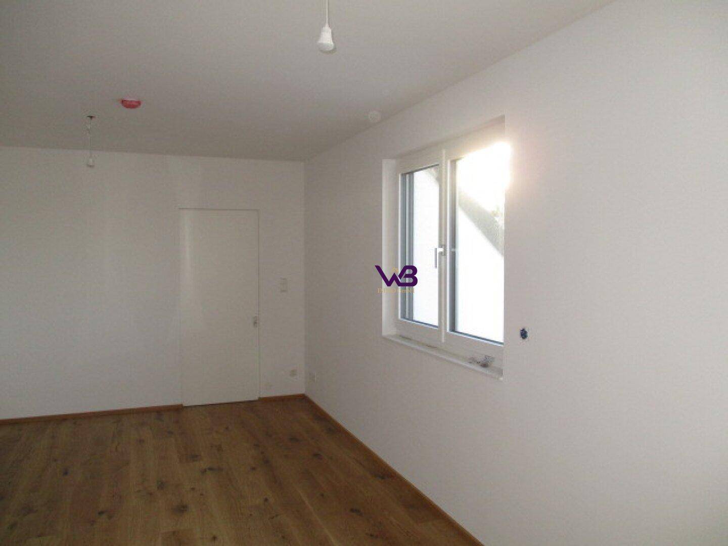Leerer Raum mit Holzboden und einem großen Fenster, das viel Sonnenlicht hereinlässt.