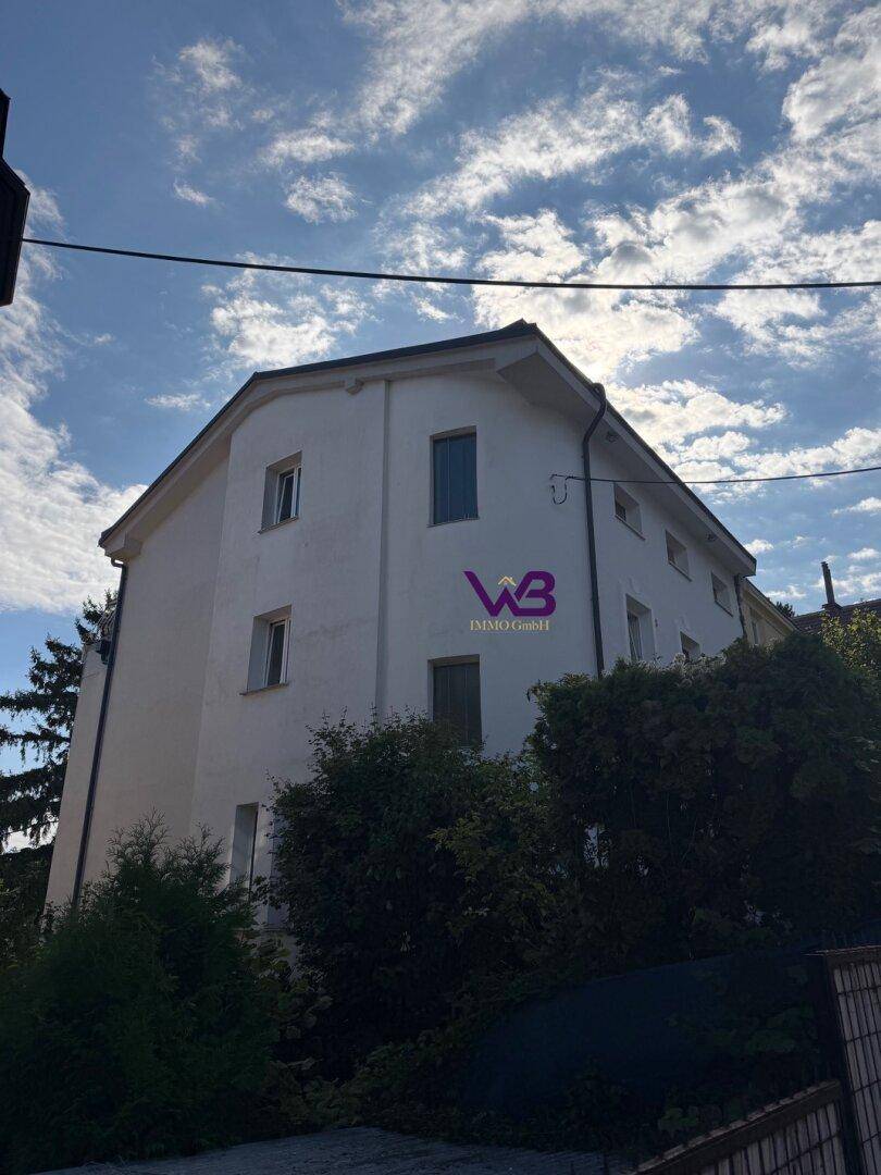 Seitenansicht des weißen Gebäudes, umgeben von üppigem Grün und hohen Bäumen.