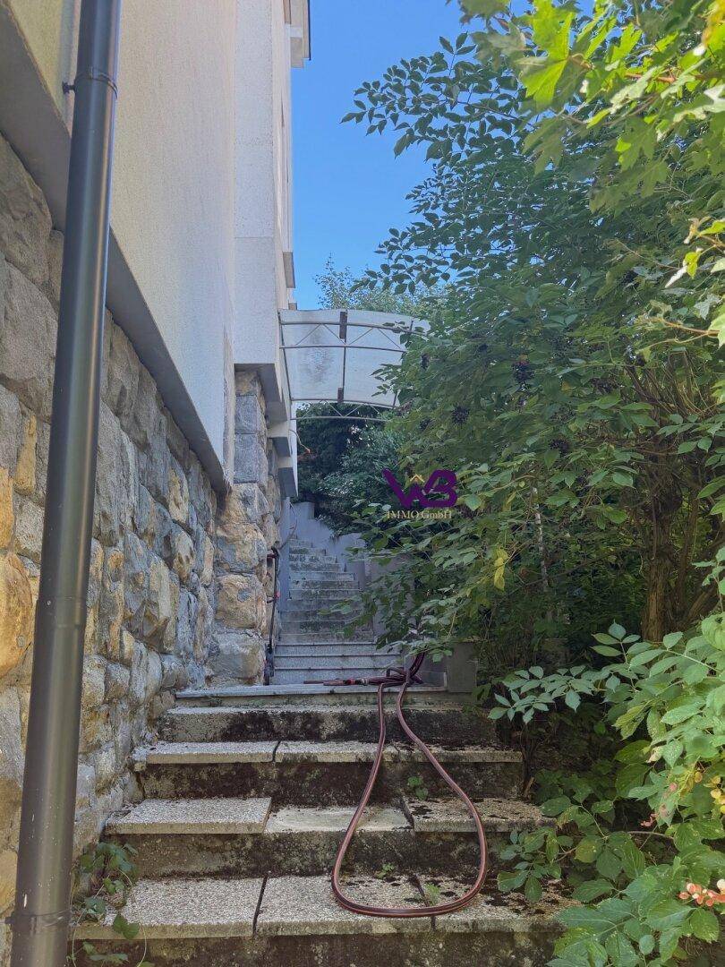 Steintreppe führt entlang der steinverkleideten Hauswand durch üppiges Grün.