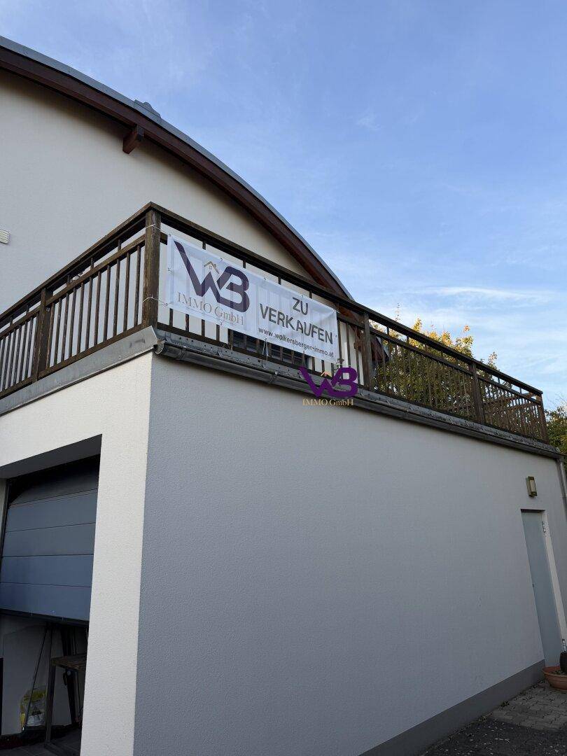 Balkon mit Holzgeländer und einem "Zu Verkaufen"-Schild, über einer Garageneinfahrt.