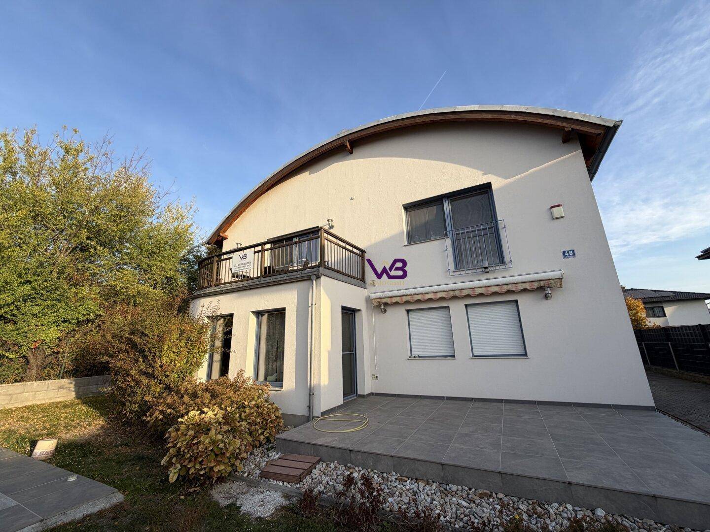 Modernes Haus mit geschwungener Dachlinie, Balkon und gepflasterter Terrasse im Vorgarten.
