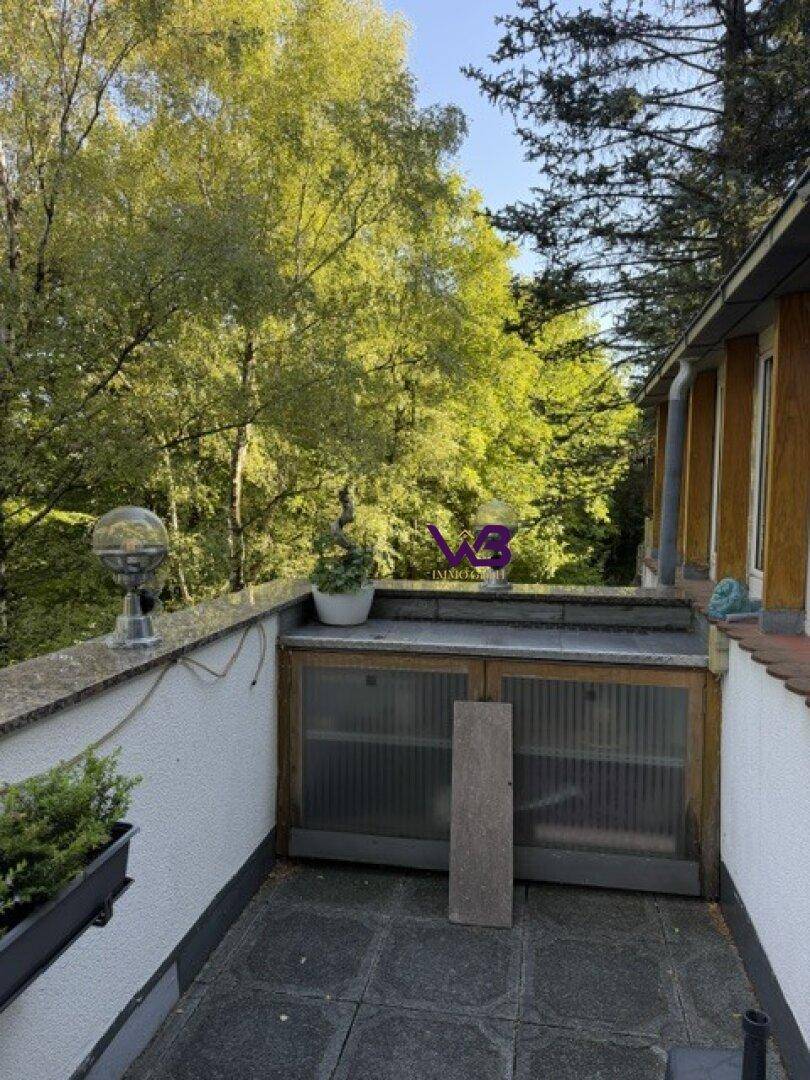 Kleine Terrasse mit gefliestem Boden, Pflanzen und Blick auf die bewaldete Umgebung.
