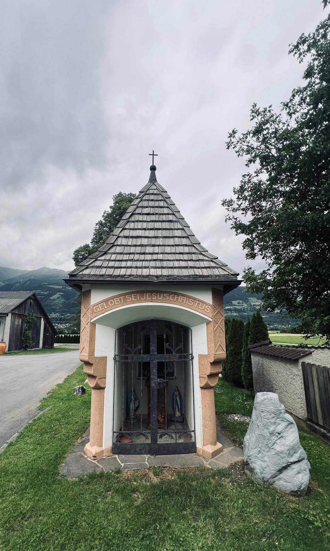 Kleiner, kunstvoll gestalteter Schrein mit Schindeldach und religiösen Figuren in ländlicher Umgebung.