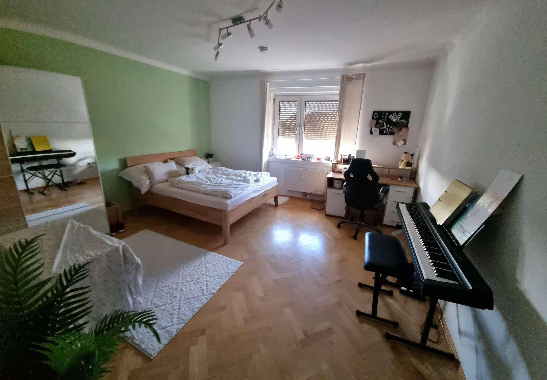 Helles Schlafzimmer mit Holzbett, grün gestrichener Akzentwand und einem Arbeitsbereich mit Musikinstrument.