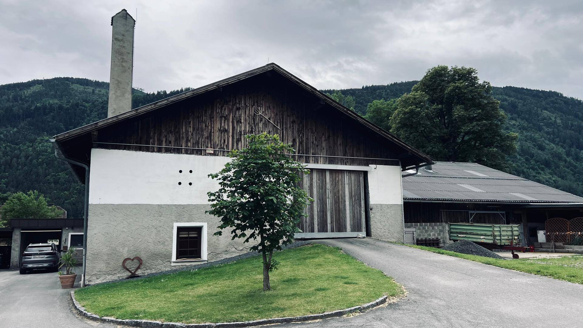 Traditionelles Scheunengebäude mit hohem Schornstein und Holzelementen, umgeben von grüner Landschaft.