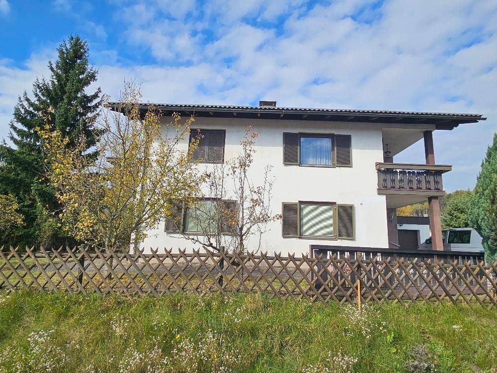 Hausfassade mit Holzzaun im Vordergrund, umgeben von Bäumen und ländlicher Umgebung.