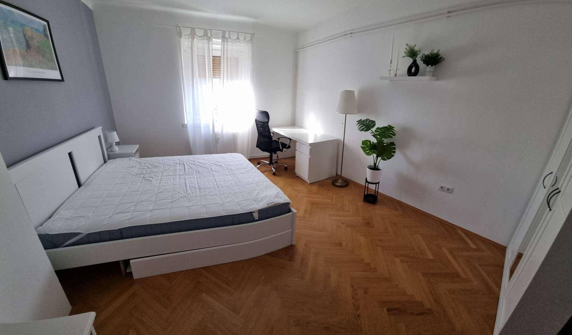Modernes Schlafzimmer mit weißem Bett, grauer Akzentwand, Schreibtisch und viel Tageslicht.