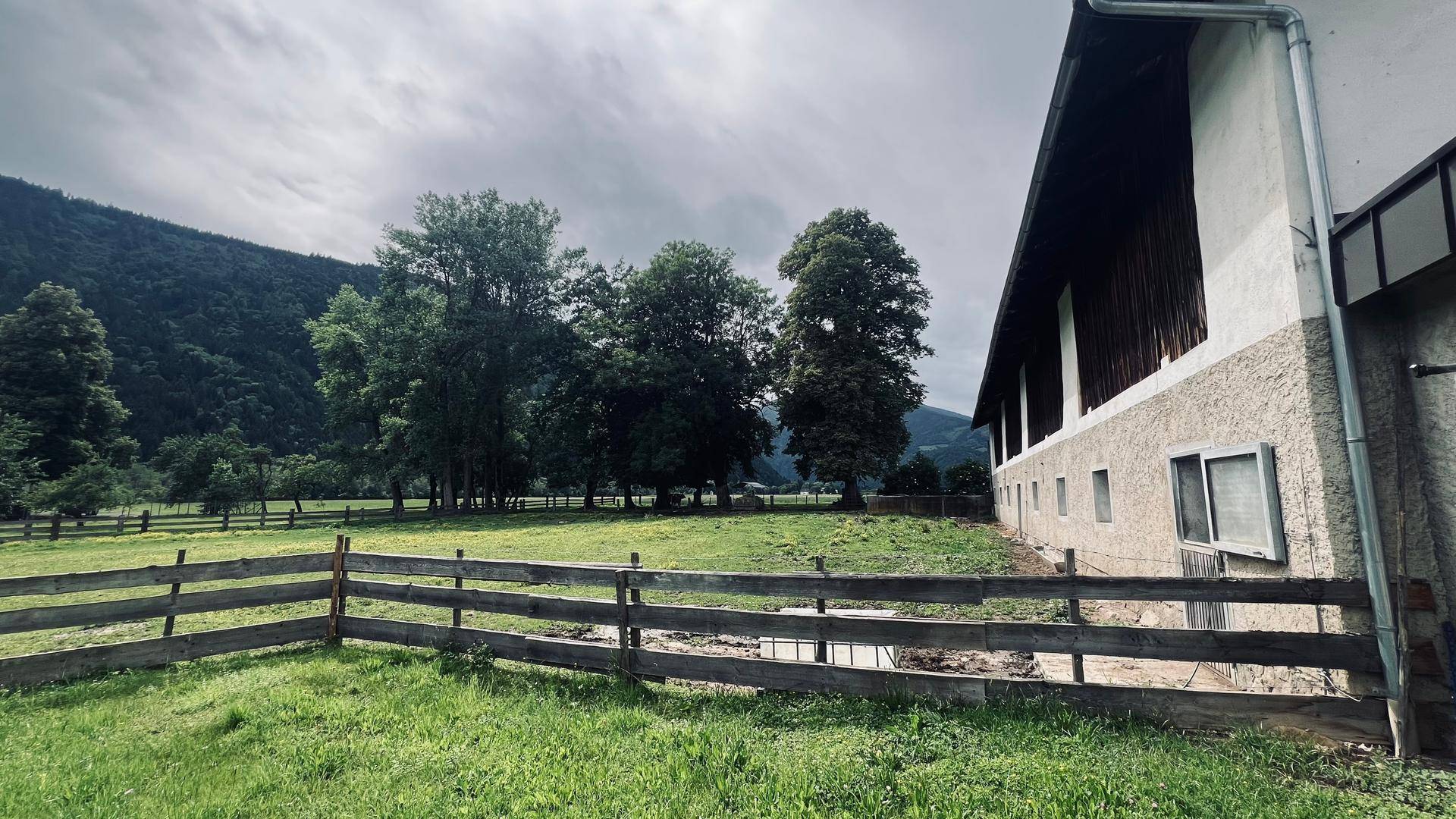 Weitläufige grüne Wiese mit Holzzaun und einem Gebäude im Hintergrund, umgeben von Bäumen.