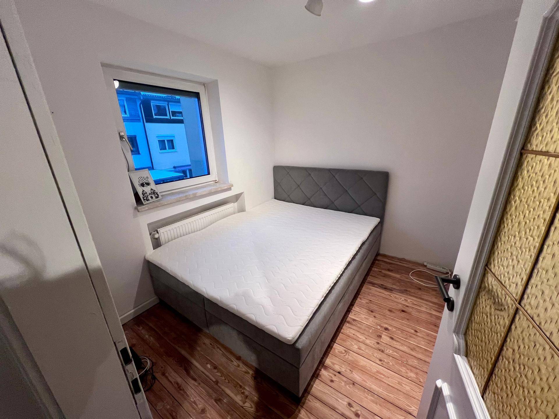 Schlafzimmer mit Doppelbett, Holzboden und Fenster, das viel Tageslicht hereinlässt.